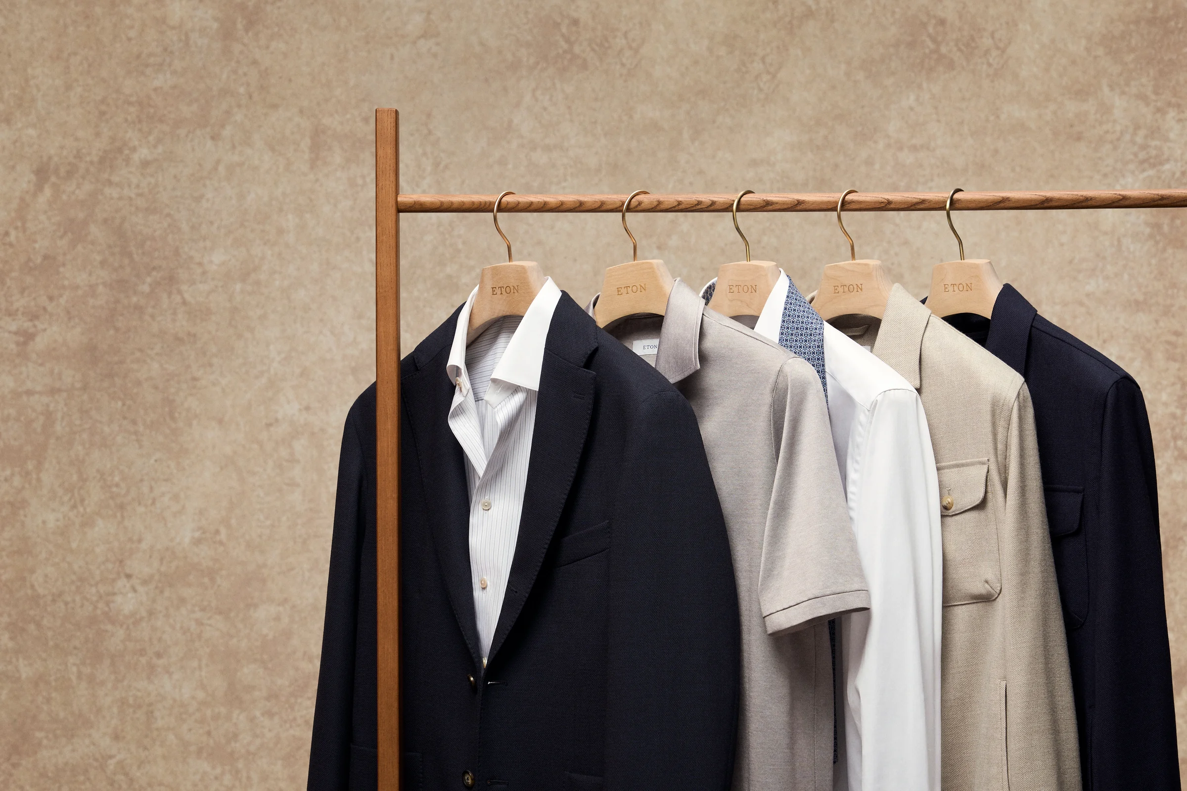 Neutral and navy Eton jackets and shirts arranged on a clothing rail against a warm stone wall